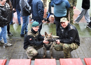 Funkcjonariusz SG i uczeń klasy mundurowej głaszczą psa służbowego SG w tle inni uczestnicy pikniku 23.04.24 r.