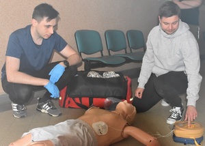 Dwóch uczestników szkolenia podczas ćwiczeń z defibrylacji na fantomie leżącym na podłodze