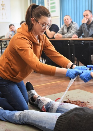 Uczestniczka szkolenia podczas ćwiczeń zakłada opatrunek na kończynę dolną. W tle uczestnicy szkolenia