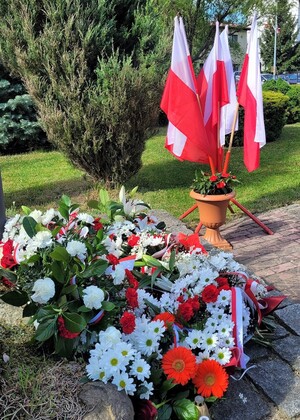Kwiaty złożone przed krzyżem w tle flagi państwowe.