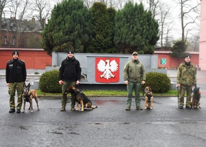 Zdjęcie grupowe Funkcjonariuszy SG z psami służbowymi Czterech Funkcjonariuszy SG stoją na tle trybuny Ośrodka. Przy nodze każdego funkcjonariusza siedzi pies służbowy.